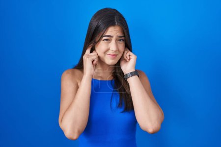 Foto de Hispanic woman standing over blue background covering ears with fingers with annoyed expression for the noise of loud music. deaf concept. - Imagen libre de derechos