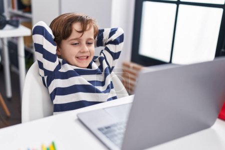 Téléchargez les photos : Adorable élève hispanique garçon utilisant un ordinateur détendu avec les mains sur la tête en classe - en image libre de droit