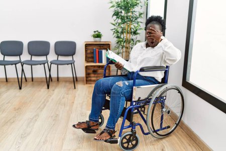 Téléchargez les photos : Young black woman sitting on wheelchair at waiting room peeking in shock covering face and eyes with hand, looking through fingers with embarrassed expression. - en image libre de droit
