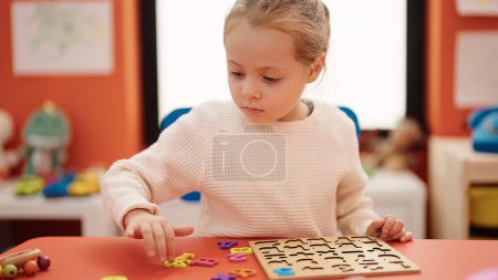 Foto de Adorable chica rubia jugando con matemáticas juego de puzzle sentado en la mesa en el jardín de infantes - Imagen libre de derechos