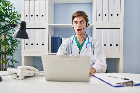 Foto de Young doctor man working on online appointment scared and amazed with open mouth for surprise, disbelief face - Imagen libre de derechos