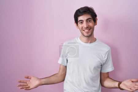 Téléchargez les photos : Jeune homme hispanique debout sur fond rose souriant gai avec les bras ouverts comme accueil amical, salutations positives et confiantes - en image libre de droit