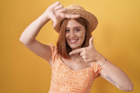 Téléchargez les photos : Jeune femme rousse debout sur fond jaune portant chapeau d'été souriant faisant cadre avec les mains et les doigts avec le visage heureux. concept de créativité et photographie. - en image libre de droit