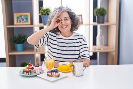 Foto de Mujer de mediana edad con el pelo gris comiendo pasteles y tomando café para el desayuno sonriendo feliz haciendo signo bien con la mano en el ojo mirando a través de los dedos - Imagen libre de derechos