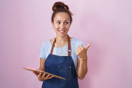 Foto de Brunette woman wearing professional waitress apron holding clipboard pointing thumb up to the side smiling happy with open mouth - Imagen libre de derechos