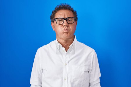 Téléchargez les photos : Middle age hispanic man standing over blue background puffing cheeks with funny face. mouth inflated with air, crazy expression. - en image libre de droit