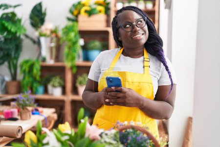 Téléchargez les photos : Afro-américaine femme fleuriste souriant confiant en utilisant smartphone chez le fleuriste - en image libre de droit