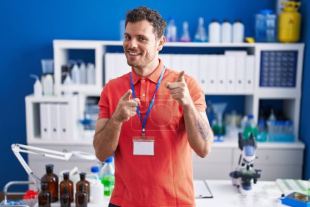 Foto de Young hispanic man working at scientist laboratory pointing fingers to camera with happy and funny face. good energy and vibes. - Imagen libre de derechos