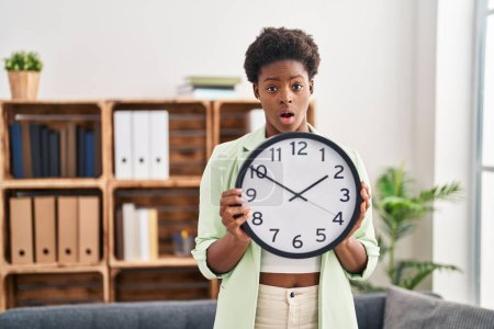Téléchargez les photos : African american woman holding big clock in shock face, looking skeptical and sarcastic, surprised with open mouth - en image libre de droit
