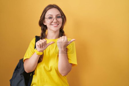Foto de Young caucasian woman wearing student backpack over yellow background pointing to the back behind with hand and thumbs up, smiling confident - Imagen libre de derechos