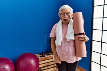 Photo for Senior woman with grey hair holding yoga mat scared and amazed with open mouth for surprise, disbelief face - Royalty Free Image