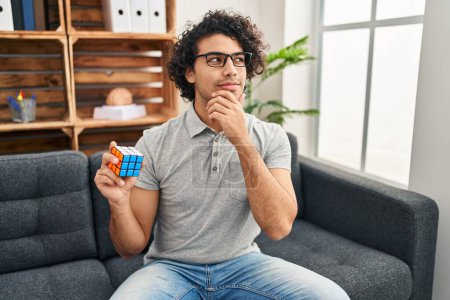 Photo for Hispanic man with curly hair playing colorful puzzle cube intelligence game serious face thinking about question with hand on chin, thoughtful about confusing idea - Royalty Free Image