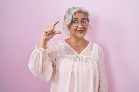 Foto de Middle age woman with grey hair standing over pink background smiling and confident gesturing with hand doing small size sign with fingers looking and the camera. measure concept. - Imagen libre de derechos