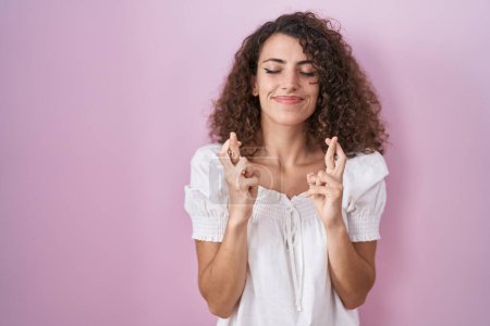 Photo for Hispanic woman with curly hair standing over pink background gesturing finger crossed smiling with hope and eyes closed. luck and superstitious concept. - Royalty Free Image
