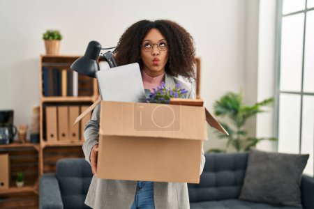 Foto de Young african american woman moving to a new office holding box with items making fish face with mouth and squinting eyes, crazy and comical. - Imagen libre de derechos