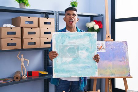 Photo for Young hispanic man holding canvas at artist studio angry and mad screaming frustrated and furious, shouting with anger looking up. - Royalty Free Image