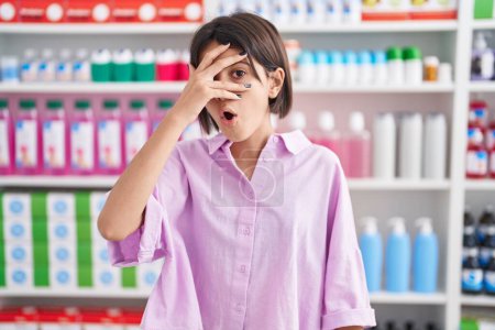 Photo for Young girl at pharmacy drugstore peeking in shock covering face and eyes with hand, looking through fingers with embarrassed expression. - Royalty Free Image