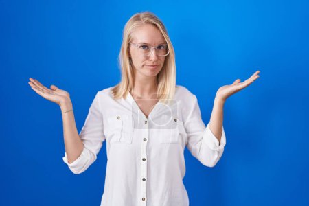 Foto de Young caucasian woman standing over blue background clueless and confused expression with arms and hands raised. doubt concept. - Imagen libre de derechos
