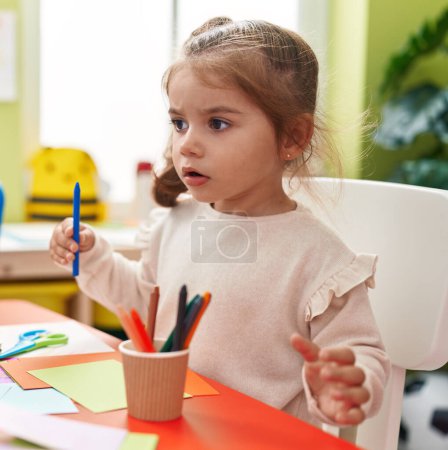 Photo for Adorable hispanic girl student sitting on table drawing on paper at kindergarten - Royalty Free Image