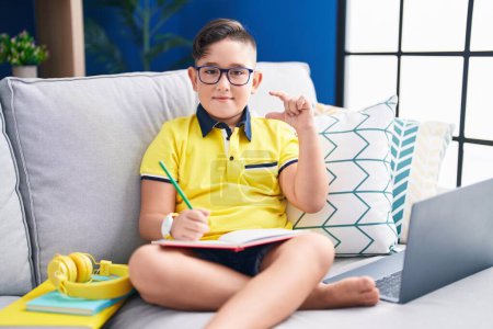 Foto de Young hispanic kid doing homework sitting on the sofa smiling and confident gesturing with hand doing small size sign with fingers looking and the camera. measure concept. - Imagen libre de derechos