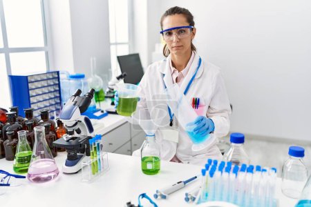 Téléchargez les photos : Young hispanic woman working at scientist laboratory relaxed with serious expression on face. simple and natural looking at the camera. - en image libre de droit