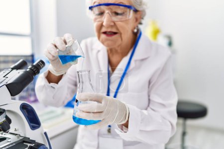 Téléchargez les photos : Femme aux cheveux gris âgée portant l'uniforme de scientifique versant du liquide au laboratoire - en image libre de droit