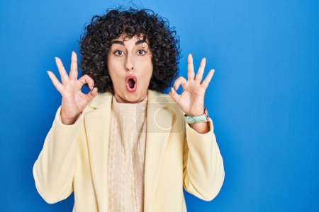 Foto de Young brunette woman with curly hair standing over blue background looking surprised and shocked doing ok approval symbol with fingers. crazy expression - Imagen libre de derechos