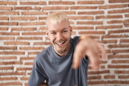 Photo for Young caucasian man standing over bricks wall laughing at you, pointing finger to the camera with hand over body, shame expression - Royalty Free Image