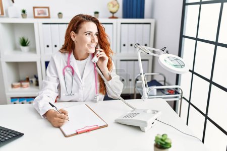 Téléchargez les photos : Jeune rousse femme portant l'uniforme de médecin parlant au téléphone à l'hôpital - en image libre de droit