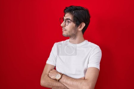 Téléchargez les photos : Young hispanic man standing over red background looking to the side with arms crossed convinced and confident - en image libre de droit