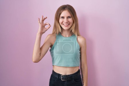 Foto de Blonde caucasian woman standing over pink background smiling positive doing ok sign with hand and fingers. successful expression. - Imagen libre de derechos