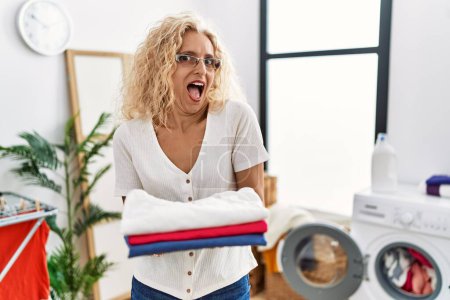 Photo for Middle age blonde woman holding folded laundry after ironing celebrating crazy and amazed for success with open eyes screaming excited. - Royalty Free Image