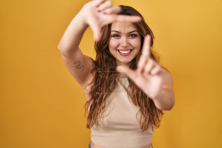 Téléchargez les photos : Young hispanic woman standing over yellow background smiling making frame with hands and fingers with happy face. creativity and photography concept. - en image libre de droit