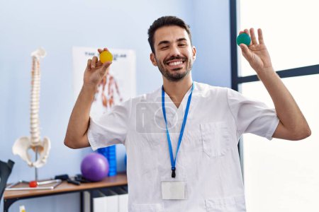 Téléchargez les photos : Young hispanic physiotherapist man holding strength balls to train hand muscles winking looking at the camera with sexy expression, cheerful and happy face. - en image libre de droit