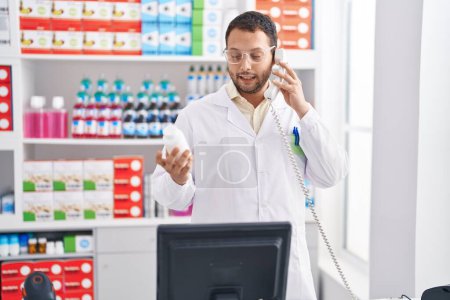 Foto de Joven farmacéutico sosteniendo pastillas botella hablando en el teléfono inteligente en la farmacia - Imagen libre de derechos