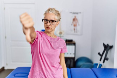 Photo for Middle age blonde woman at pain recovery clinic looking unhappy and angry showing rejection and negative with thumbs down gesture. bad expression. - Royalty Free Image
