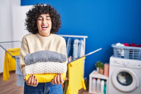 Téléchargez les photos : Young brunette woman with curly hair holding clean laundry smiling and laughing hard out loud because funny crazy joke. - en image libre de droit