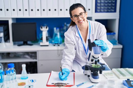 Téléchargez les photos : Young hispanic woman scientist write on document using microscope at laboratory - en image libre de droit
