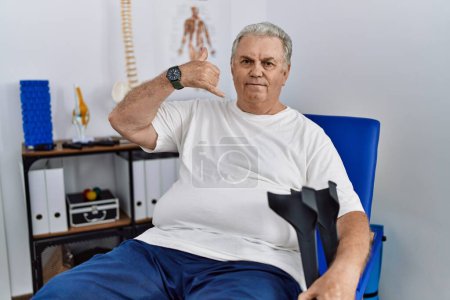 Foto de Senior caucasian man at physiotherapy clinic holding crutches smiling doing phone gesture with hand and fingers like talking on the telephone. communicating concepts. - Imagen libre de derechos