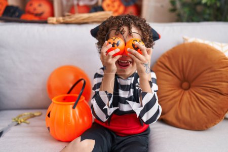 Foto de Adorable hispanic boy having halloween party holding pumpkin baskets over eyes at home - Imagen libre de derechos
