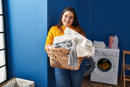 Photo for Young beautiful plus size woman smiling confident holding basket with clothes at laundry room - Royalty Free Image