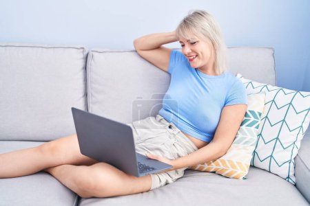 Foto de Mujer rubia joven usando el ordenador portátil sentado en la mesa en casa - Imagen libre de derechos