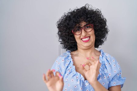 Foto de Young brunette woman with curly hair wearing glasses over isolated background disgusted expression, displeased and fearful doing disgust face because aversion reaction. with hands raised - Imagen libre de derechos