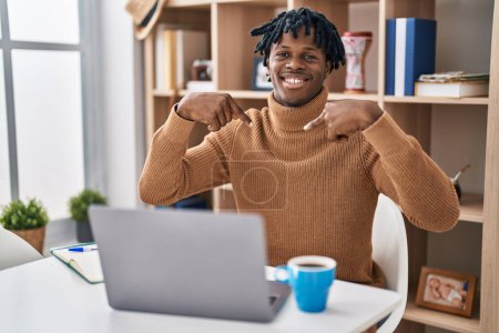 Téléchargez les photos : Jeune homme africain avec dreadlocks travaillant à l'aide d'un ordinateur portable à l'air confiant avec le sourire sur le visage, se montrant du doigt fier et heureux. - en image libre de droit