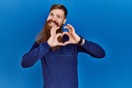 Photo for Redhead man with long beard wearing casual blue sweater over blue background smiling in love doing heart symbol shape with hands. romantic concept. - Royalty Free Image