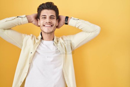 Foto de Joven hombre hispano de pie sobre fondo amarillo relajante y estiramiento, brazos y manos detrás de la cabeza y el cuello sonriendo feliz - Imagen libre de derechos