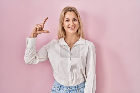 Téléchargez les photos : Young caucasian woman wearing casual white shirt over pink background smiling and confident gesturing with hand doing small size sign with fingers looking and the camera. measure concept. - en image libre de droit