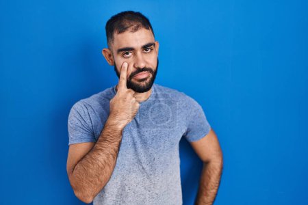 Téléchargez les photos : Moyen-Orient homme avec la barbe debout sur fond bleu pointant vers l'oeil vous regarder geste, expression suspecte - en image libre de droit