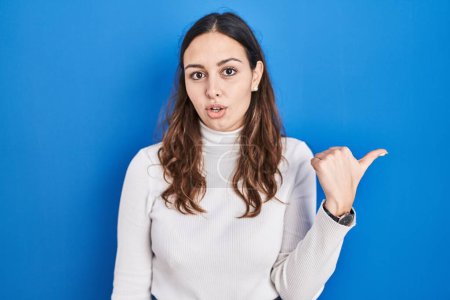 Photo for Young hispanic woman standing over blue background surprised pointing with hand finger to the side, open mouth amazed expression. - Royalty Free Image