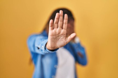 Téléchargez les photos : Femme afro-américaine avec des tresses debout sur fond jaune couvrant les yeux avec les mains et faisant arrêter geste avec expression triste et peur. concept embarrassé et négatif. - en image libre de droit